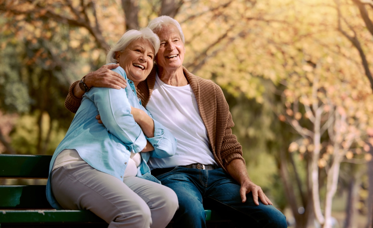 An elderly couple sits on a park bench, smiling and embracing, with autumn trees in the background.