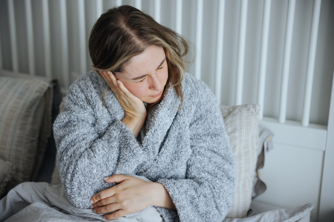 A person in a fluffy gray robe sits on a bed, holding their head with one hand and their arm with the other, appearing uncomfortable or unwell.